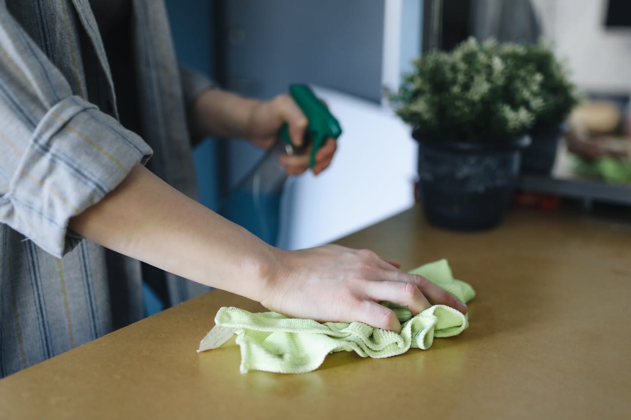 services-04 Close-up of a woman wiping a table with a spray bottle and cloth indoors, symbolizing effective housekeeping.